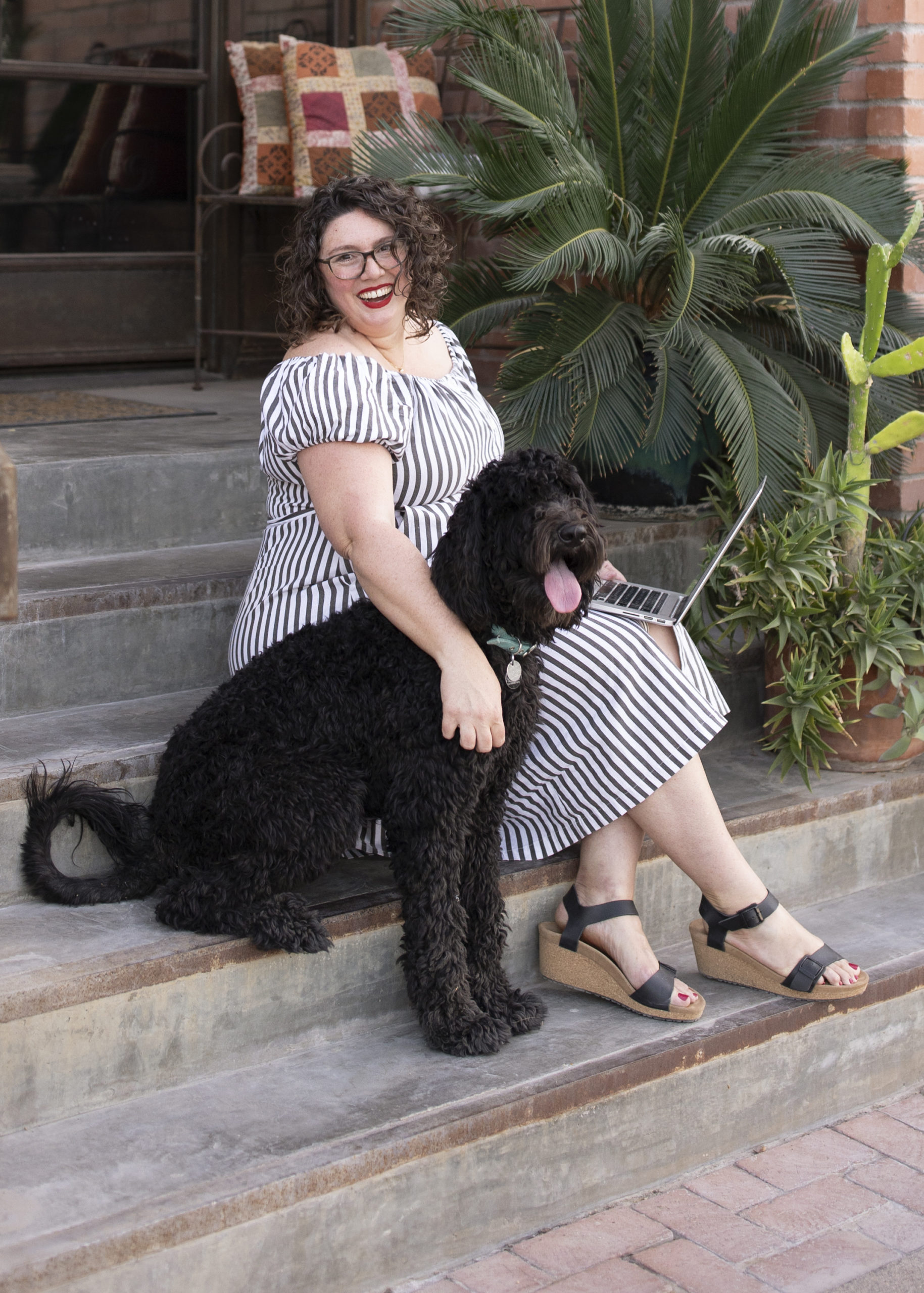 Woman sits on a step with an open laptop and her labradoodle beside her.